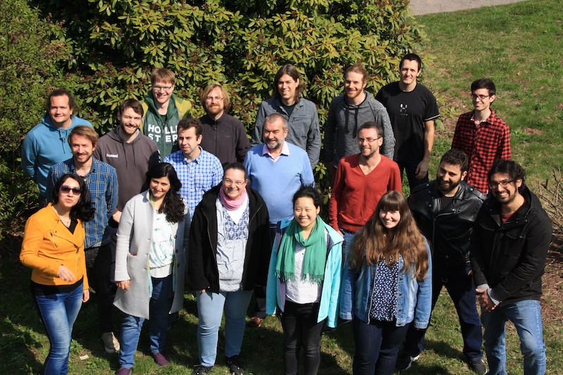 Group of 19 people standing on grass next to some shrubs, smiling upward at the camera.