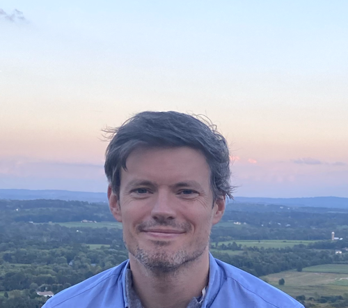 Smiling man in blue shirt with farmland, trees and and blue sky behind him.