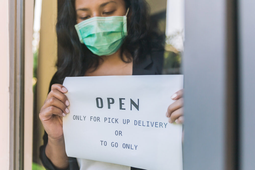 A Latina essential worker wearing a face mask posts a sign on a restaurant window saying "open only for pickup delivery or to go" during the pandemic
