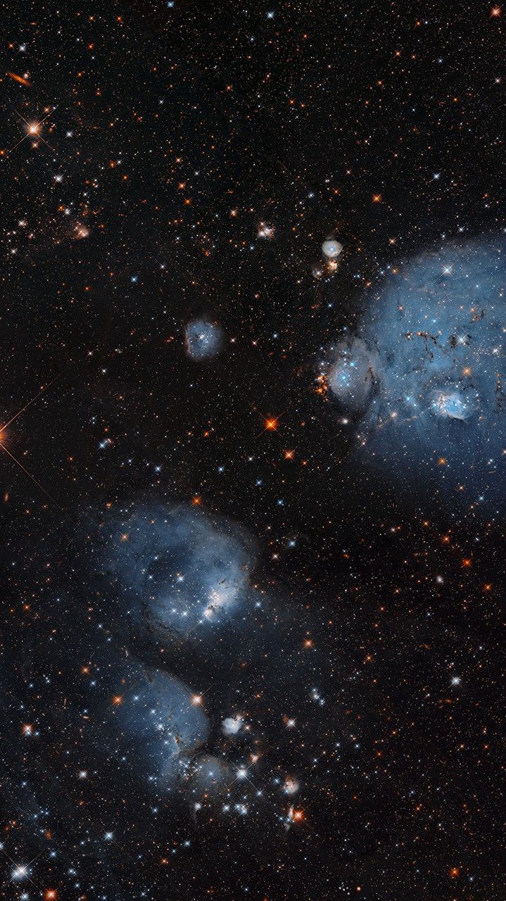 Bluish-white, bubble-shaped clouds of gas dominate this image, sprinkled with clusters of stars. Plentiful stars and some background galaxies are visible throughout the image. The gas clouds are concentrated in the upper right and lower middle of the image.
