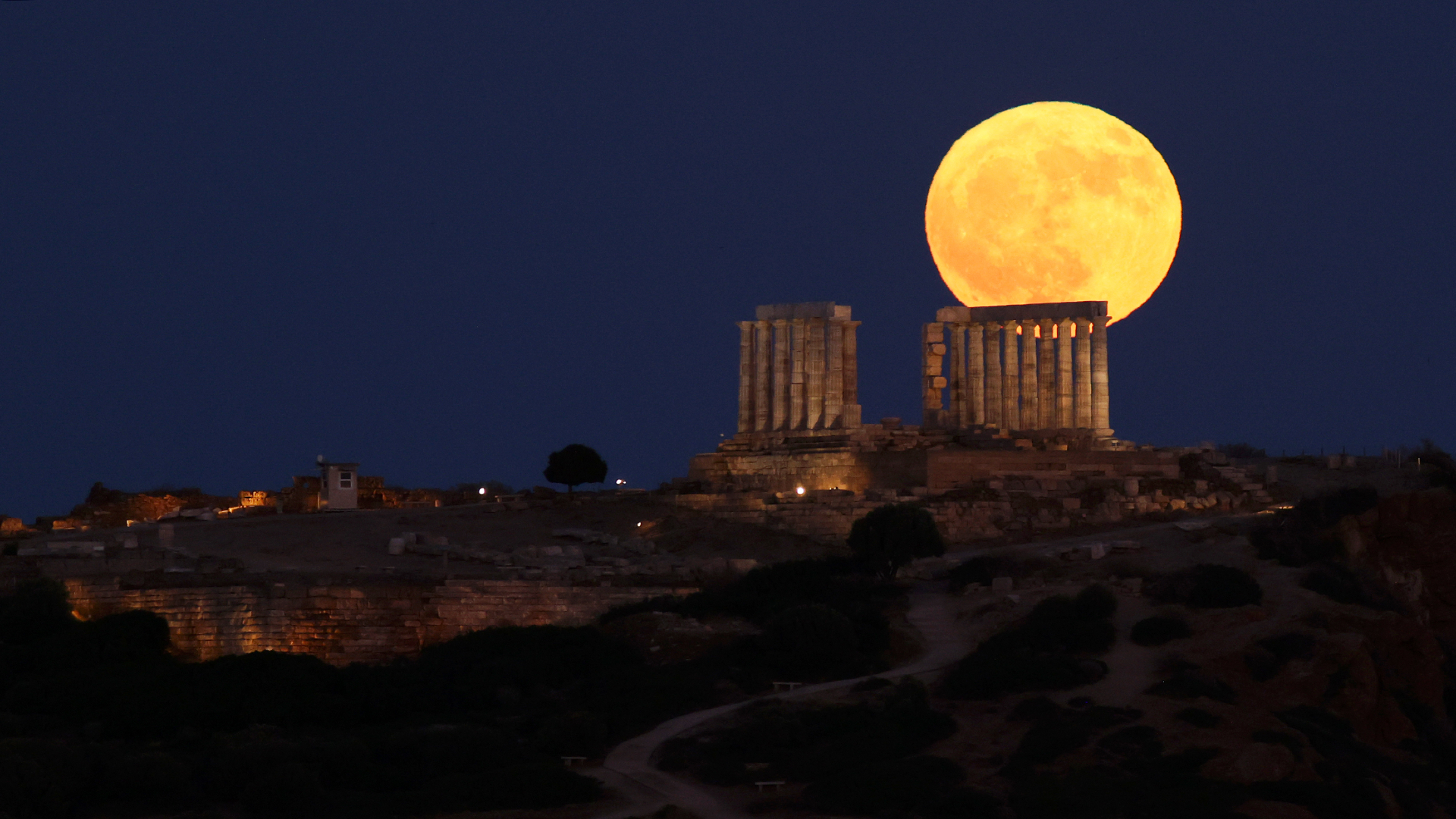 An orange full moon is pictured rising over an ancient columned temple ruin on a hillside.