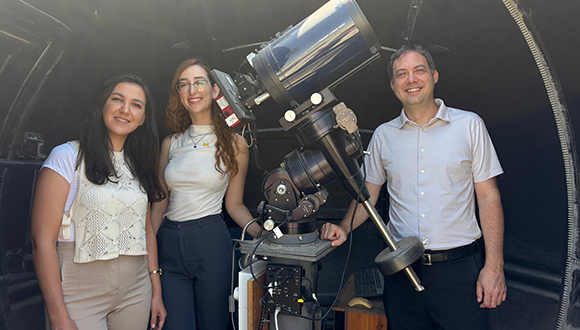 Two young women with long hair at left, a telescope and a middle aged man at right smiling for the camera.