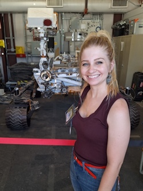Smiling woman wearing a tank top and standing in front of a model of a Mars rover.