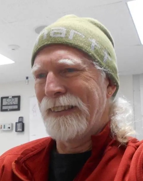 Smiling man with white beard, moustache and pony tail, wearing a red jacket.
