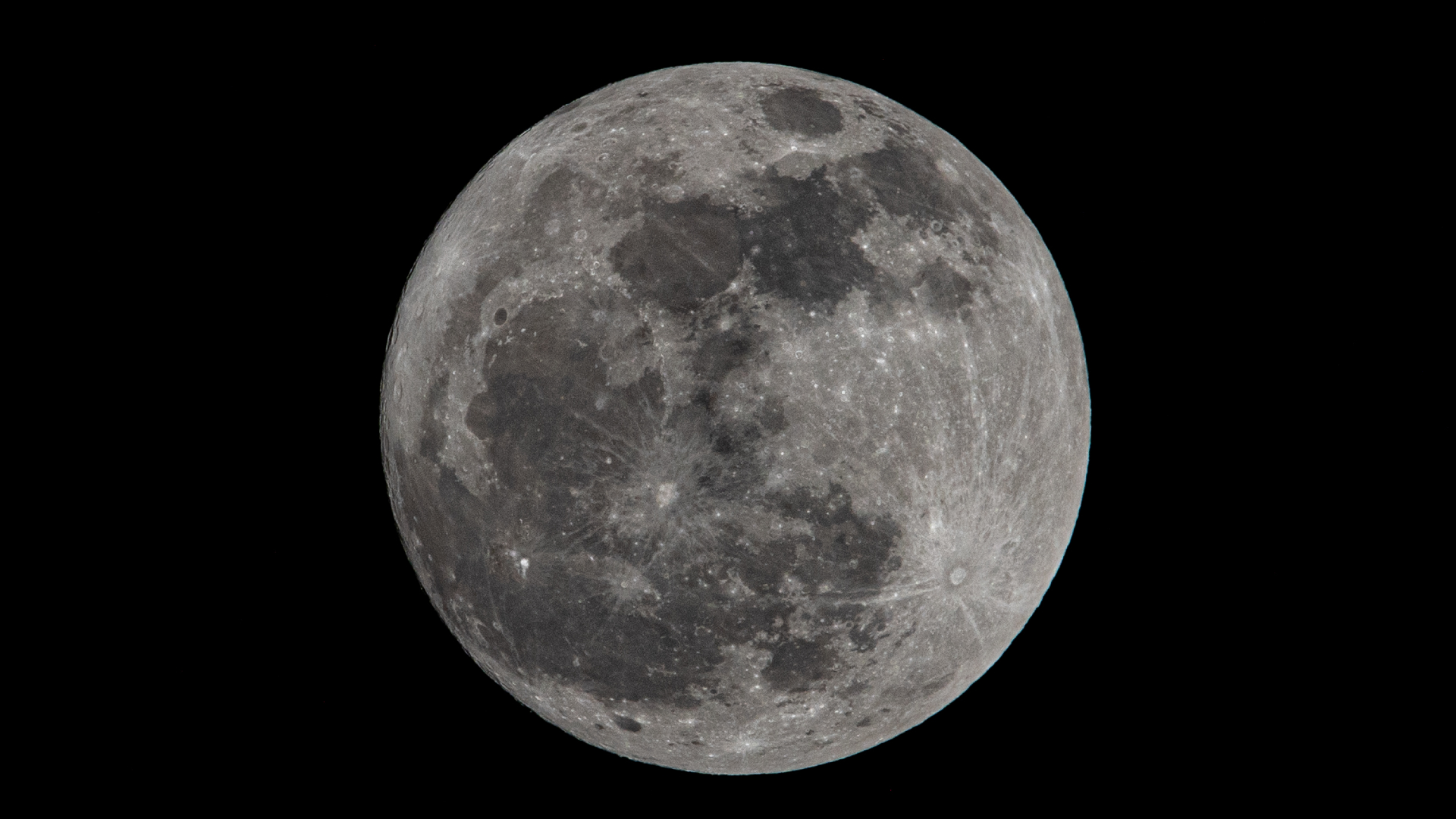 A detailed picture of a full moon against a dark sky. Dark basaltic regions called lunar maria, or lunar seas, can be seen marking swathes of the surface, along with myriad craters and bright spots of material thrown out by ancient impacts.