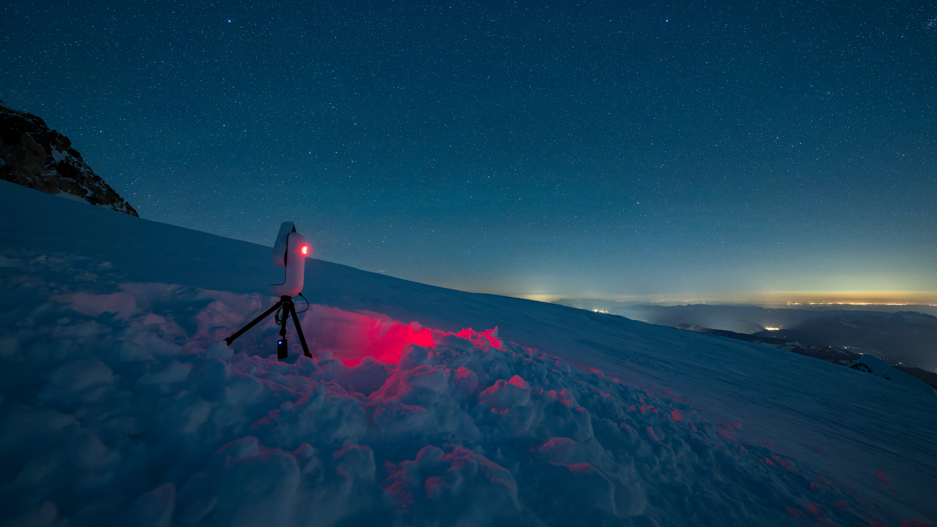 A white smart telescope is pictured bathing the nearby mountain snow with red light at night. Stars can be seen in the sky overhead, while artificial light glows on the horizon below.