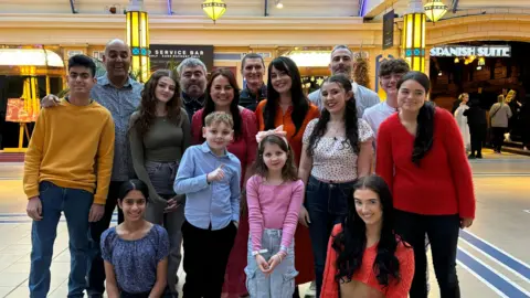 Natt Morris A group of 15 people, across a range of ages, who make up part of the stammering choir. They are all smiling at camera in a lobby area.