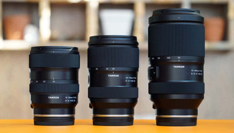 Three black Tamron camera lenses of varying sizes are displayed upright on an orange surface with a softly blurred background.