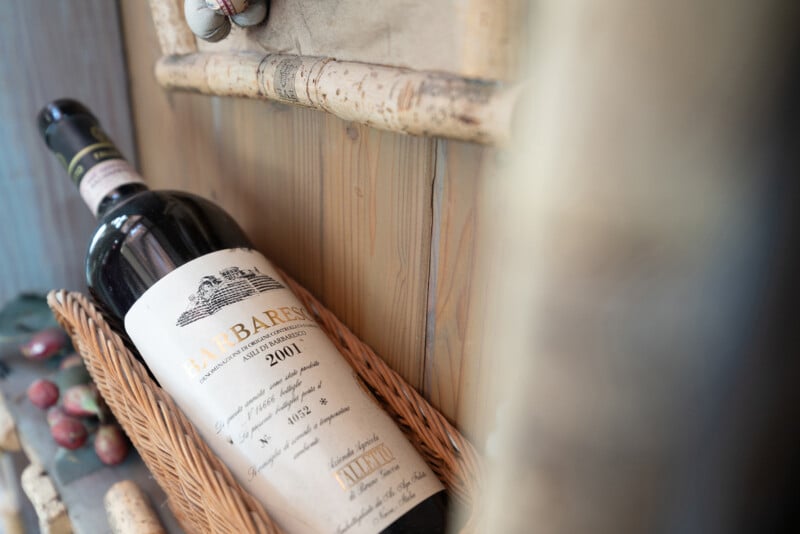 A close-up of a Barbaresco 2001 wine bottle lying in a wicker basket, with wine corks and a baseball bat in the background on a wooden surface.