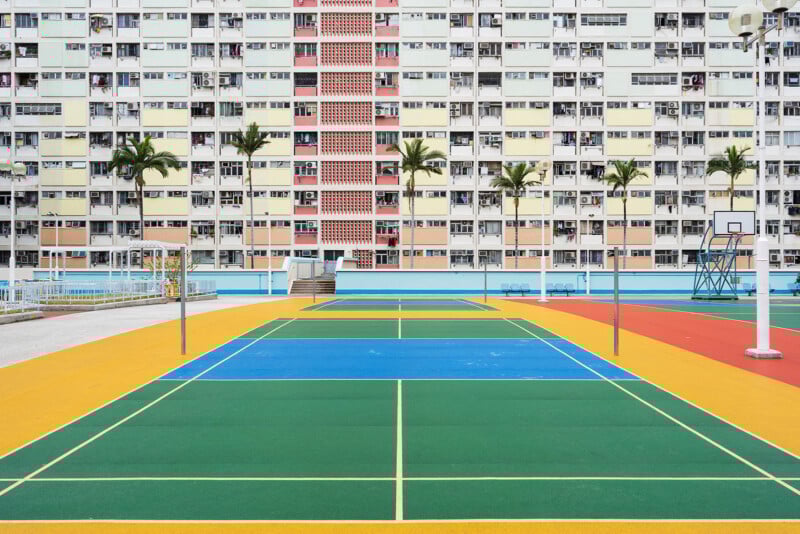 A colorful outdoor sports court with green, blue, and yellow sections, palm trees, and a tall residential building with multicolored balconies in the background.
