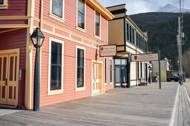 A wooden boardwalk runs past colorful historic buildings with visitor center signs in a small mountain town, with a snow-capped peak visible in the background.