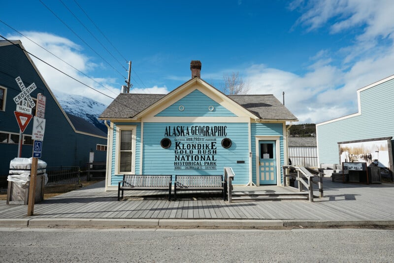 A small blue wooden building with a sign reading "Alaska Geographic Klondike Gold Rush National Historical Park" stands on a wooden boardwalk under a partly cloudy sky. Benches and wooden railings are in front.
