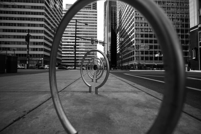 Metal bike racks form concentric circles along a city sidewalk, with tall office buildings and traffic lights in the background, captured in black and white.