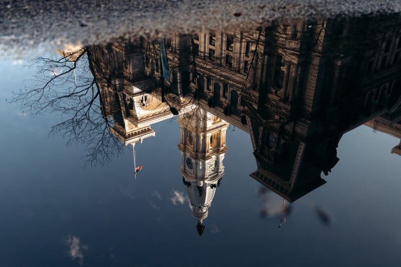 A historic building with a clock tower and a U.S. flag is reflected in a puddle on the ground, with bare tree branches visible to one side. The image is inverted due to the reflection.