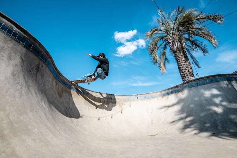 A skateboarder performs a trick on the edge of an empty concrete pool under a bright blue sky, with a palm tree in the background.