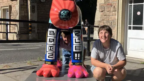 Teenagers Leo and Hugh crouch beneath a large, colourful Gromit sculpture in the same way they did as children. The sculpture is covered in bright designs and patterns. The boys smile.