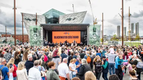 Belfast Tradfest The grey and glass triangular Titanic Musuem sticks out the back of a stage and orange screen with big writing that says 'BLACKWATER CÉILÍ BAND' on it. Hundreds of people are in front of the stage, standing and some are dancing.