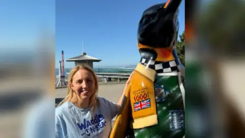 Elliot Deady/BBC Amy Dearing is standing to the left of the image with her arms resting on a penguin sculpture on the right. She has shoulder length blonde hair and is wearing a blue T-shirt that says Waddle-on-Sea on the front.
