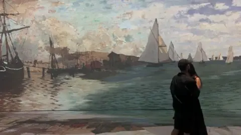Jermaine Foster/BBC A young couple dressed in black stand to view a projection of one of Claude Monet's paintings, in which sailing ships are depicted in a harbour.
