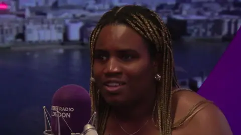 BBC Esther Obigbesan speaks into a BBC Radio London microphone in a studio, with the Thames and London skyline visible on the backdrop behind her.