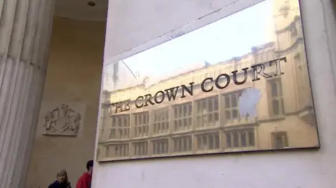 BBC A brass plaque on a sandstone building reading "Crown Court". A column and a court crest is visible in the background. Two people can be seen walking into the building.