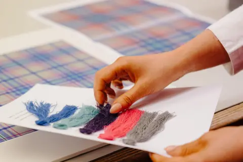 Glasgow 2026 A close up of the designer's hands, touching the thread swatches over the tartan design.