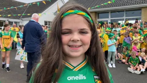 Molly is smiling, wearing her green and gold Donegal jersey. She has a green and gold headband in her brown hair. She is standing in front of a large crowd of children, all dressed in Donegal colours.