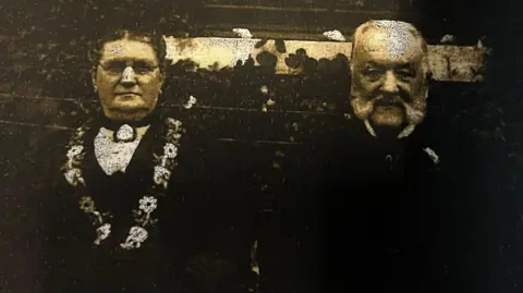 Archive A man and woman, of late middle age, dressed in Victorian clothing. The black and white picture is very faded.