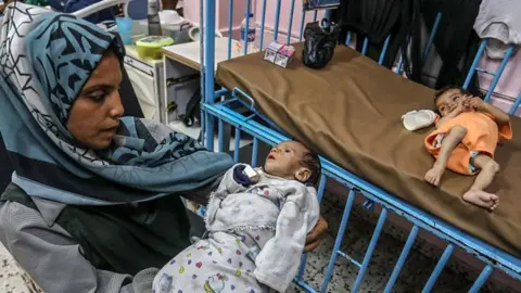 BBC Woman holding an apparently malnourished child as another one lies in a bed
