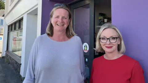 BBC Jennifer Merrett, left, is a woman wearing a grey bowl-neck jumper. She is smiling and has blue eyes and fair hair. She has sunglasses on her head and is stood outside the purple walls of the salon. Racheal Day, right, is a petite woman with short bobbed blonde hair, black glasses and red lipstick. She is stood smiling and wearing a red dress.