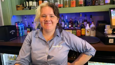Meg wearing a blue branded shirt at the Philharmonic bar. She has blonde and black hair split down the middle and is wearing hoop earrings with several piercings around her nose. There are bottles of alcohol behind her at the bar with a fridge on her right.