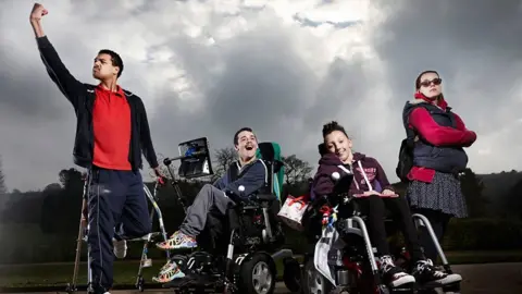 BBC Four people outside, with one person on the left standing using sticks and raising a fist. Two others are seated in wheelchairs, one of which has a communication device attached, and another is stood with their arms crossed and wearing sunglasses. A cloudy sky and trees are visible in the background.