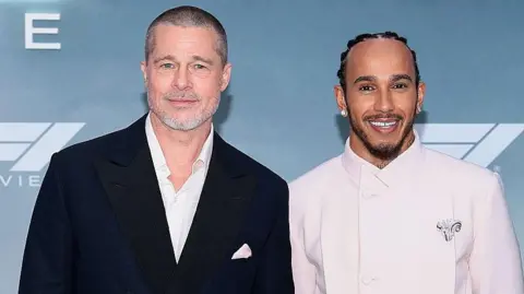 Getty Images Brad Pitt, with a shaven head and grey beard, is stood next to Lewis Hamilton in front of branding for the F1 movie