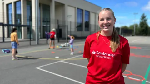 Mimi is wearing a red 'Santander International' t-shirt stands on an outdoor sports court. In the background, other people are participating in activities, and a building labeled 'Sports Centre' is visible under a clear blue sky