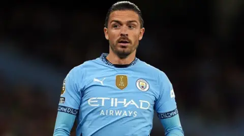 PA Media Footballer Jack Grealish runs on a football pitch. He is wearing a sky blue Manchester City jersey with long sleeves underneath it. 