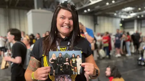 BBC A woman is wearing a black t-shirt that says 'Back to the Beginning'. She is holding a photograph with the Osbourne family and smiling.