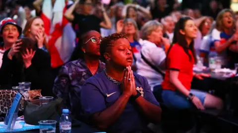 Getty Images A woman in a blue England T-shirt holds her hands in the prayer position surrounded by fans in Boxpark, Croydon, London, in a particularly tense moment during the semi-final between England and Italy 