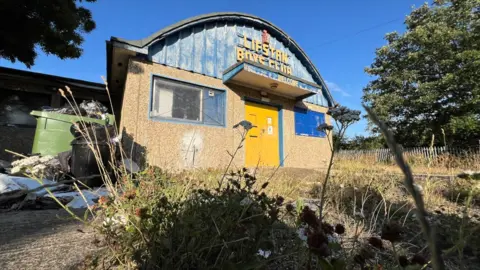 Stuart Woodward/BBC The Lifstan Boys Club building, surrounded by overgrown grass and weeds, with an overflowing green bin to the side. The building has a pebble-dashed front with two blue-framed windows and a yellow front door. The upper half of the building is a domed curved roof with blue metal fronting and the words Lifstan Boys Club in yellow writing.