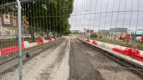 Ed Barnes A road fenced off behind metal fencing. The road surface is under construction and is lined by red and white plastic barriers.