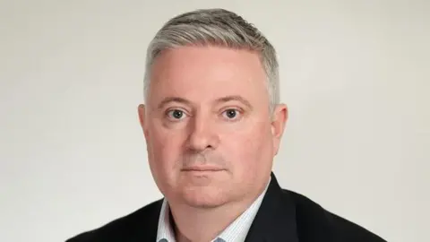 EPUT Paul Scott looking at the camera with a serious expression. He is wearing a black suit jacket and a white shirt without a tie. He has short grey hair.