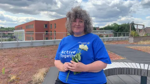 BBC A woman with grey curly hair is sitting outdoors on a bench and smiling. She is wearing a bright blue t-shirt with the words Active Together and is holding a white rose