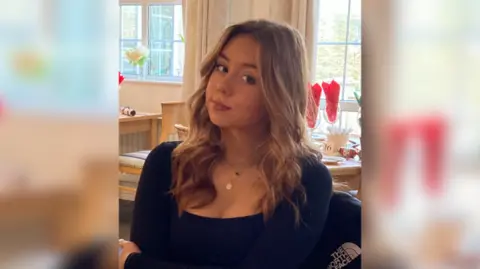 Glasser family Jessica with brown highlighted hair in loose, wavy curls. She is wearing a black square-necked top. She is sitting down in a restaurant with brown wooden tables behind her.