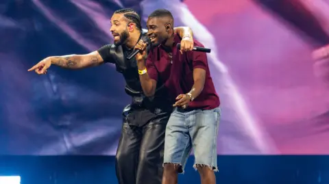 Getty Images Both artists are on the stage with Drakes hand over Dave who sings into his microphone. Drake is pointing to the crowd, he is in all black with Dave in maroon t shirt and denim shorts. 