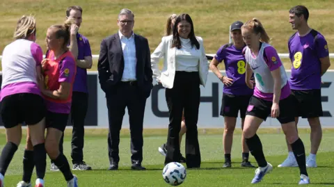 PA Media The Prime Minister, Sir Keir Starmer and The Secretary of State for Culture, Media and Sport Lisa Nandy, both dressed in suits, watch and England training session on a green pitch.