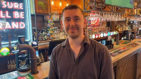 Tom stood at the bar. He is wearing a black shirt and has short dark hair. Beer taps, a shelf of spirits and glasses can be seen behind him along with neon signage.