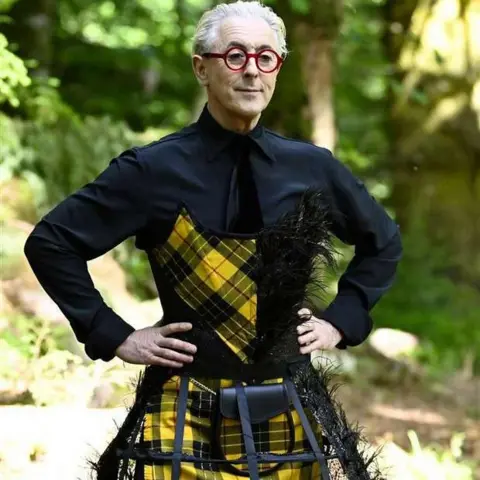 Peacock TV A snap from Traitors USA of Alan Cumming in a black shirt and a yellow and black tartan kilt and bustier.