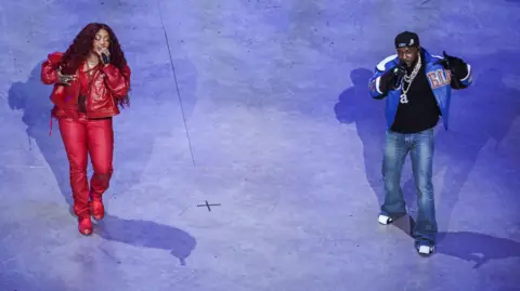 PA Media SZA, in a red leather trousers, jacket and boots and Kendrick Lamar in jeans, white trainers, a black jumper and blue bomber jacket, perform on a blue stage. The photo is taken from above.