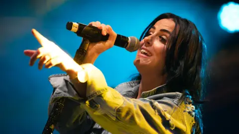 Getty Images A woman with long brown hair singing into a mic as she wears a blue denim jacket.