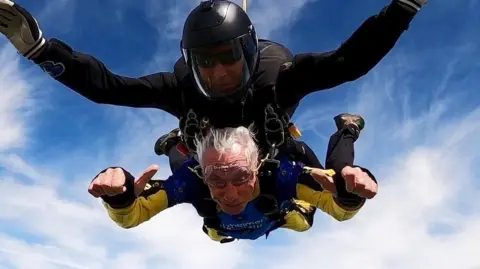 Brian Standring Brian, who has grey hair and wears a yellow and blue skydiving suit, gives two thumbs up to the camera as he skydives. He is attached in tandem to an instructor, who is wearing all black.
