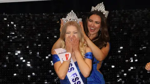 Cutter Creatives Claire Redgrave and Lauren Jennings pictured in front of a sparkly black background. Both are wearing sequined blue dresses. A smiling Lauren is putting a tiara on Claire's head, while Claire has her hands over her mouth in disbelief. Claire has long blonde hair and has a Miss Essex sash on, while Lauren has long brunette hair.